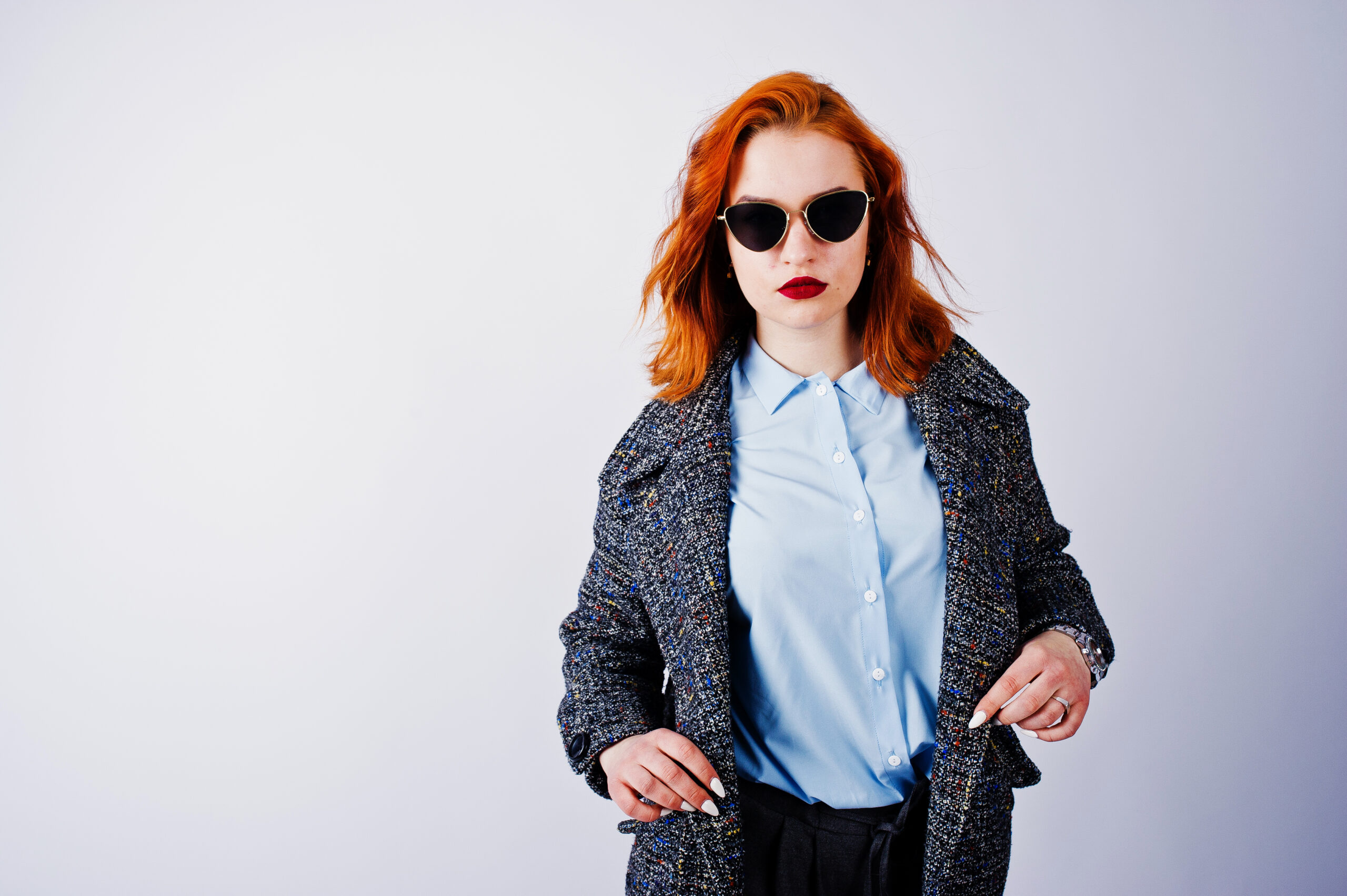 Portrait of a fantastic redheaded girl in blue shirt, grey overcoat posing with sunglasses in the studio.